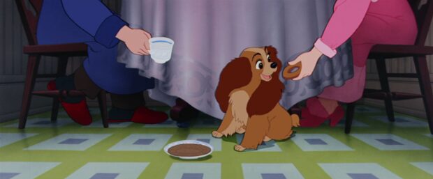 Lady’s owners feeding her at the table, seen from the dog’s perspective with only their arms visible in Lady and the Tramp (1955).